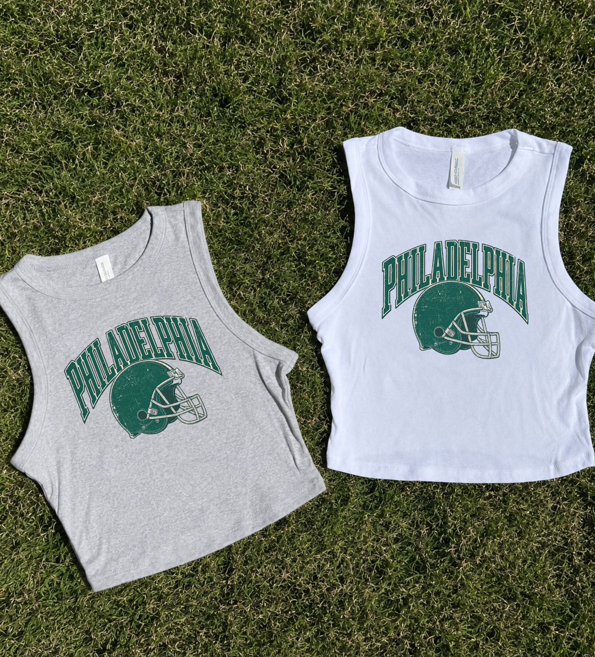 Two sleeveless cropped tank tops laid flat on green grass. The left top is light heather gray with a green Philadelphia football helmet and arched 'PHILADELPHIA' graphic.