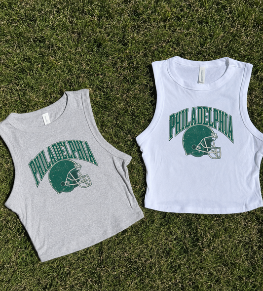Two sleeveless cropped tank tops laid flat on green grass. The left top is light heather gray with a green Philadelphia football helmet and arched 'PHILADELPHIA' graphic.